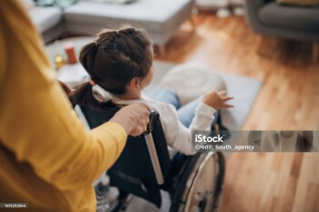 Mother pushing her young daughter who is in wheelchair in living room at home.