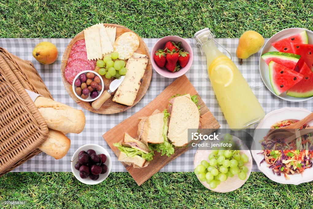 Summer picnic food scene. Top view with blanket over a green lawn background. Picnic basket, fruit, salad, sandwiches and snacks.