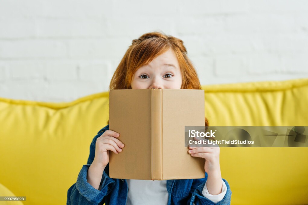 Child sitting on sofa and holding book in front of her face