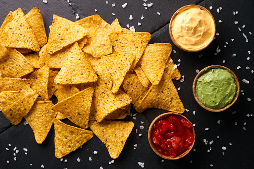 Triangular corn chips, mexican nachos with sauces tomato ketchup, cheese and guacamole in wooden bowl on dark background, close-up.