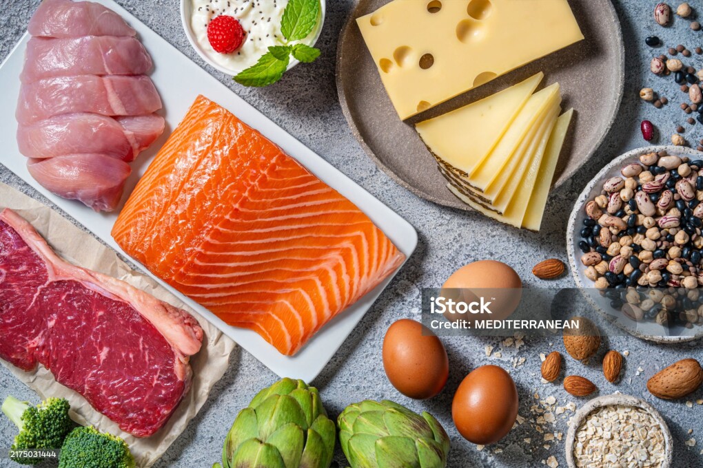 Assorted healthy protein source food good for body building ingredients as: salmon, chicken, beef, fish, eggs, beans legumes and nuts, artichoke, broccoli, cheese, yogurt, and eggs on a kitchen gray table