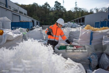 Female worker at industrial recycling site, wearing safety gear & hardhat, inspects sorted plastic pellets for reuse in manufacturing. Circular economy concept.