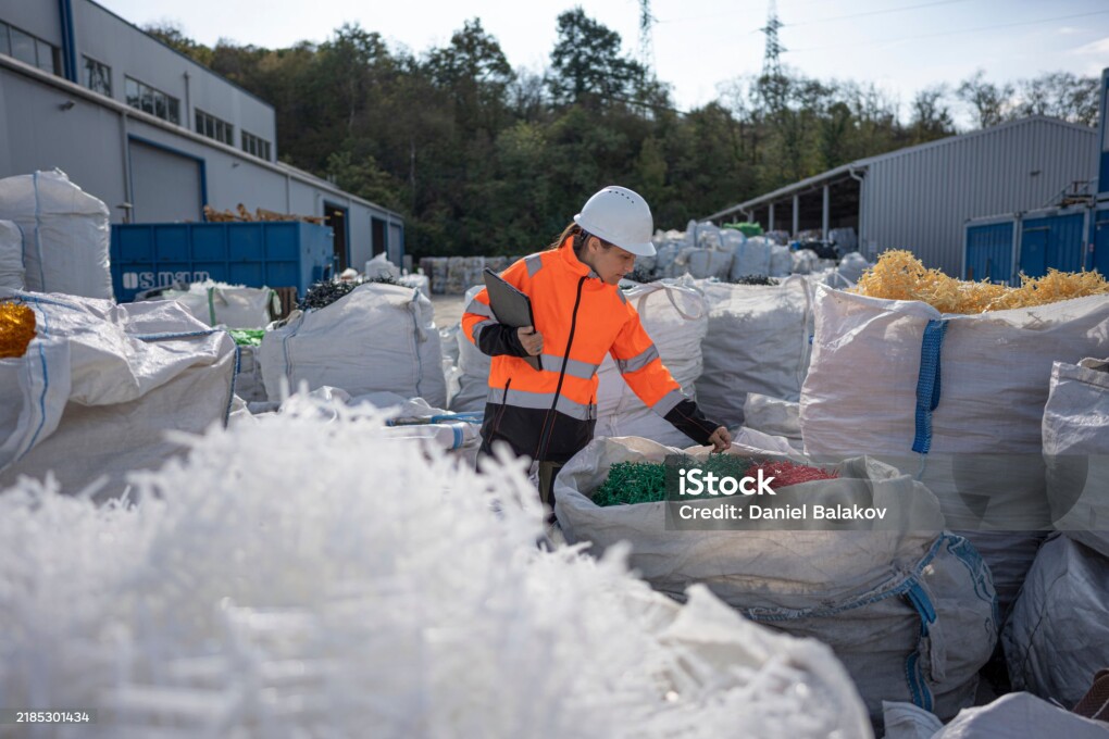 Female worker at industrial recycling site, wearing safety gear & hardhat, inspects sorted plastic pellets for reuse in manufacturing. Circular economy concept.