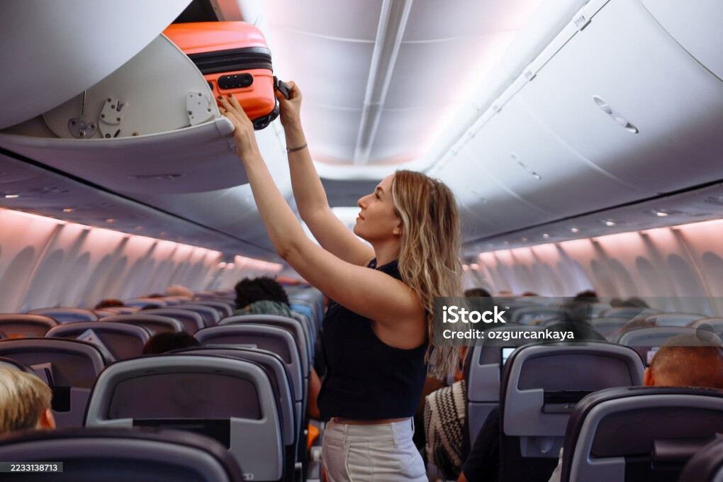 Woman travel by airplane. Passenger putting hand baggage in lockers above seats of plane. hands are seen placing or retrieving a pink hard-shell suitcase from an airplane's overhead storage compartment.