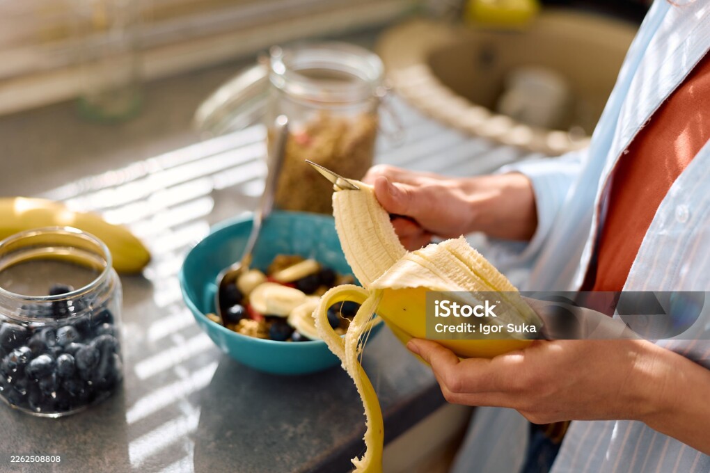 Person's hands peeling a banana over a turquoise bowl filled with berries, sliced bananas, and granola, preparing a healthy breakfast in a sunlit kitchen