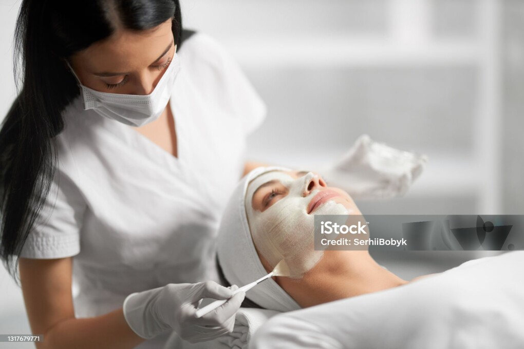 Side view of beautiful brunette beautician applying special white cream for face young cute patient. Concept of process procedure for improving face skin with cosmetics.