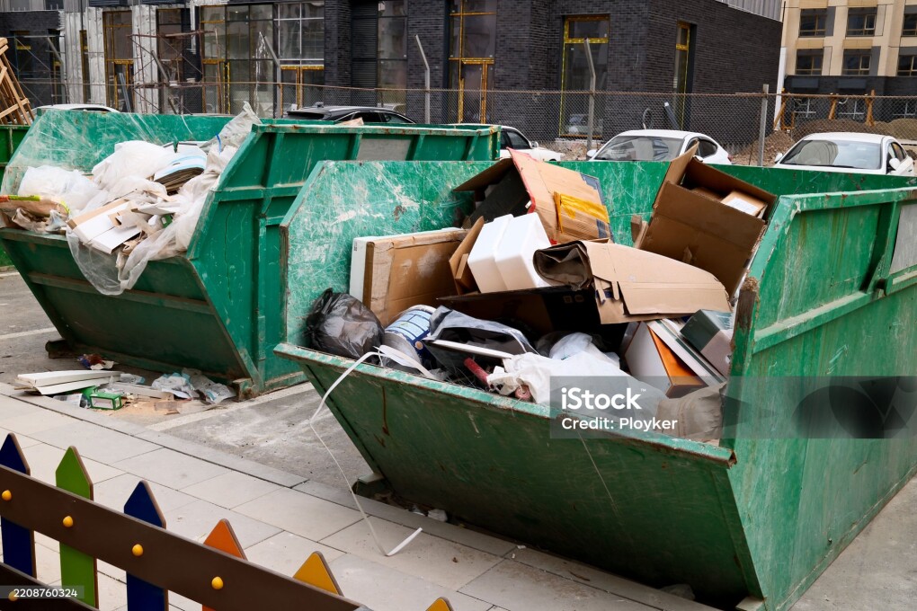 Overflowing green construction dumpster filled with cardboard boxes and building waste at urban construction site
