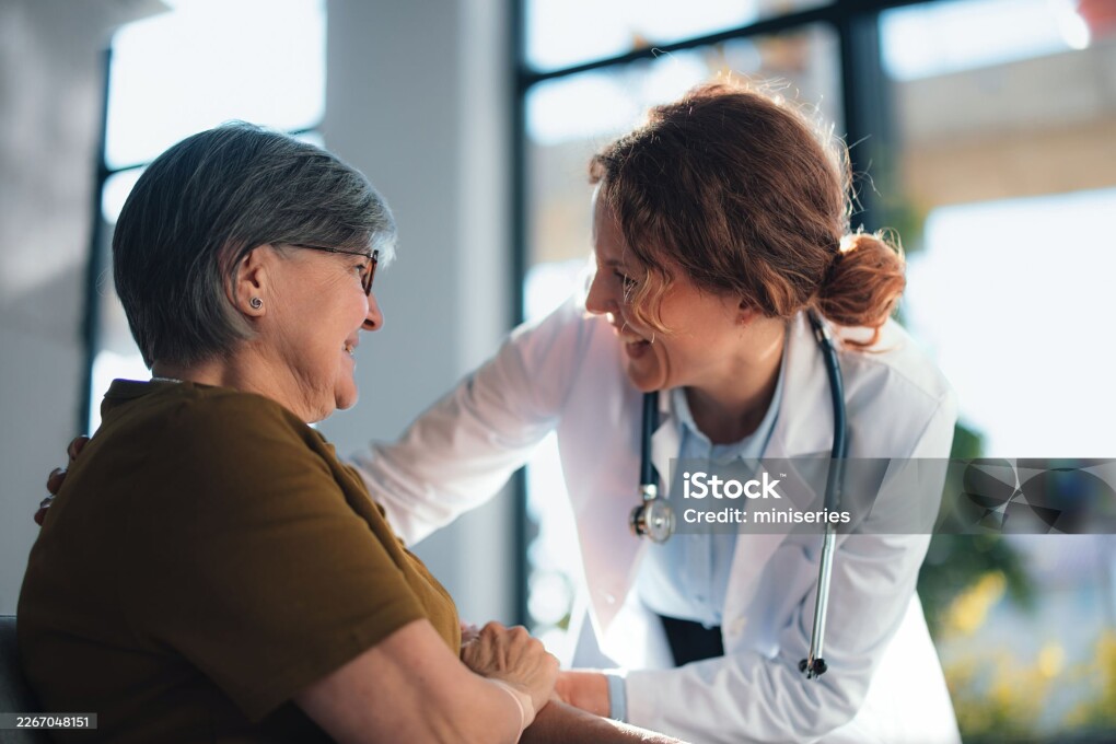 A compassionate doctor leans toward a smiling senior woman in a hospital office, offering care and reassurance during a medical consultation focused on health, trust and patient-centered care.