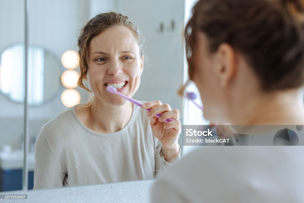 A woman brushes her teeth in front of a bathroom mirror during a calm morning routine, smiling as she practices daily oral hygiene and self-care in a bright, home environment.