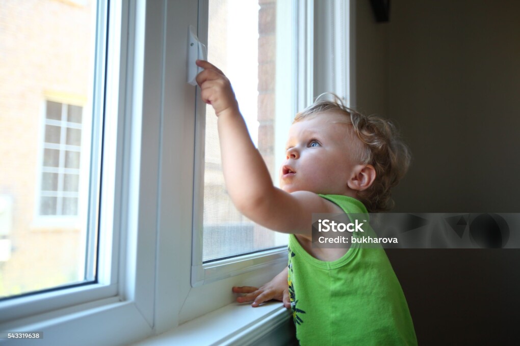 Little boy standing in front of the window and trying to open it touching the handle. Baby proofing. Danger of household items for babies and toddlers.
