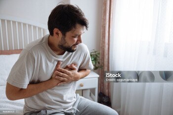 Heart attack, man with chest pain suffering at home, health problems concept. Young man with eyes closed holding his chest in discomfort, suffering from chest pain while sitting on bed at home. Elderly and health issues concept