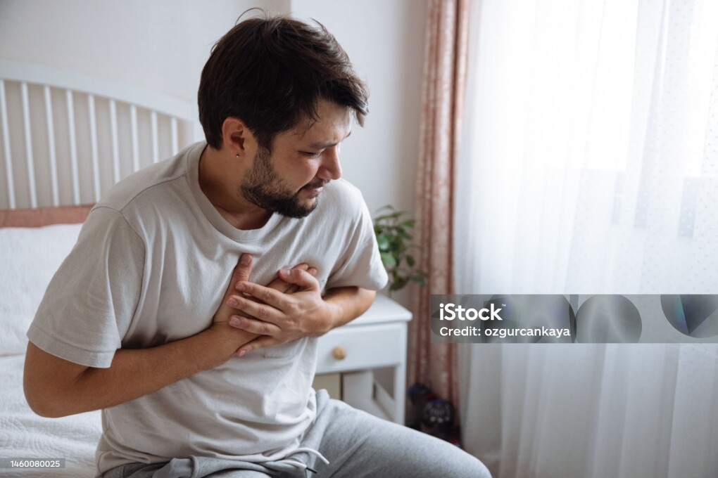 Heart attack, man with chest pain suffering at home, health problems concept. Young man with eyes closed holding his chest in discomfort, suffering from chest pain while sitting on bed at home. Elderly and health issues concept