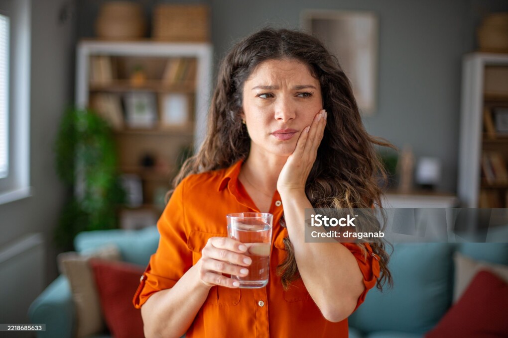 Mid adult woman holding glass of water having terrible toothache at home
