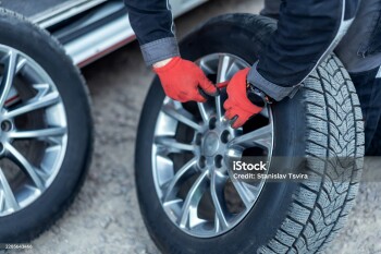 A person is working on changing the tires of a car in a garage. The individual wears gloves and uses a tool to tighten a wheel. It is late afternoon.