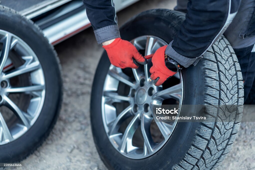 A person is working on changing the tires of a car in a garage. The individual wears gloves and uses a tool to tighten a wheel. It is late afternoon.